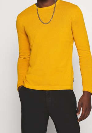 Mand iført en tekstureret gul langærmet sweater, sorte bukser og et sølvkædehalskæde, med hænderne i lommerne.