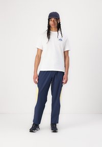 Hvit grafisk t-skjorte med blå logo, kombinert med marineblå joggebukser med gule sideakenter, og en mørkeblå caps. Svart treningssko.