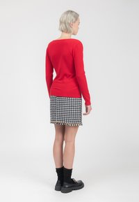 Roter Pullover mit langen Ärmeln, kombiniert mit einem schwarz-weißen Hahnentrittmuster-Rock. Das Outfit zeichnet sich durch ein strukturiertes Strickmuster und einen mit Gold akzentuierten Saum aus. Schwarze klobige Schuhe.