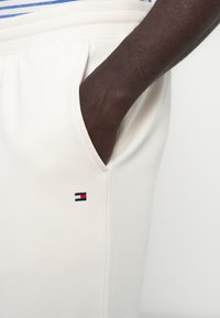 Personne à la peau foncée avec la main dans la poche portant un pantalon blanc avec un petit logo rectangulaire rouge, blanc et bleu près de la poche.