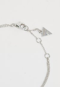 Pulsera de cadena de plata con un cierre de langosta y un charm triangular que presenta la palabra "GUESS". Los eslabones son redondos y alargados, mostrando un acabado pulido.