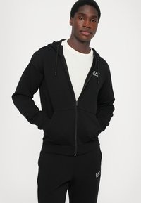 Črna zip-up hoodie s vrvicami, mehak material, sprednji žepi in izšit logotip na prsih. V kombinaciji s črnimi trenirkami.