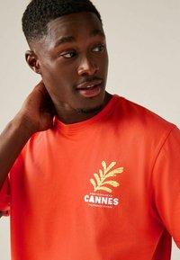 Oranž puuvillane T-särk, millel on kuldne palmilehe grafika ja tekst "Midditerranee Cannes Prantsuse Rivieras". Lühikesed varrukad ja ümar kaelus.