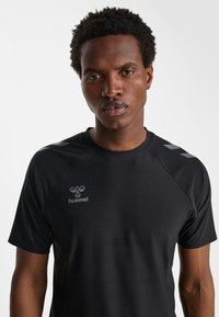 Man met kort krullend haar, gekleed in een zwart Hummel sportshirt met logo en chevron-details op de schouders, tegen een effen witte achtergrond.