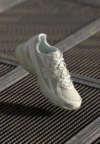 Scarpa atletica in rete beige e tan chiaro con lacci appoggiata in diagonale su una superficie di grate metalliche.