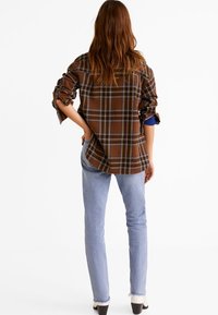 Chemise à carreaux marron avec des manches retroussées, présentant une coupe large. Associée à un jean bleu clair, coupé aux chevilles, mettant en avant des chaussures noires.