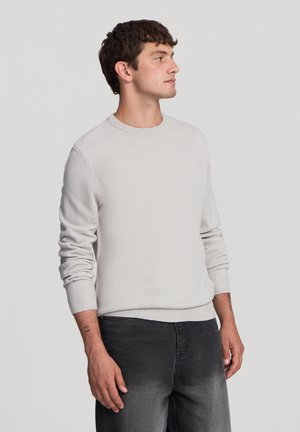 Maglione grigio chiaro realizzato in tessuto a maglia, con scollatura a girocollo e polsini a costine. Indossato con jeans grigio scuro, il modello è rivolto di lato.