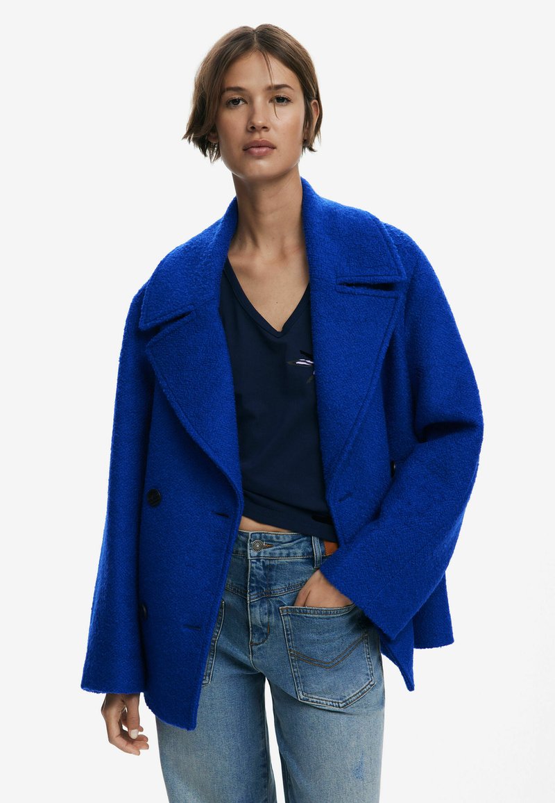 Manteau double boutonnage en laine mélangée texturée bleu vif avec de grands revers, boutons noirs et coupe décontractée, porté sur un haut marine et un jean.