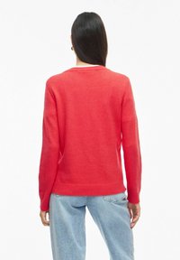 Femme aux longs cheveux foncés portant un pull corail rouge et un jean bleu clair, debout de dos à la caméra.