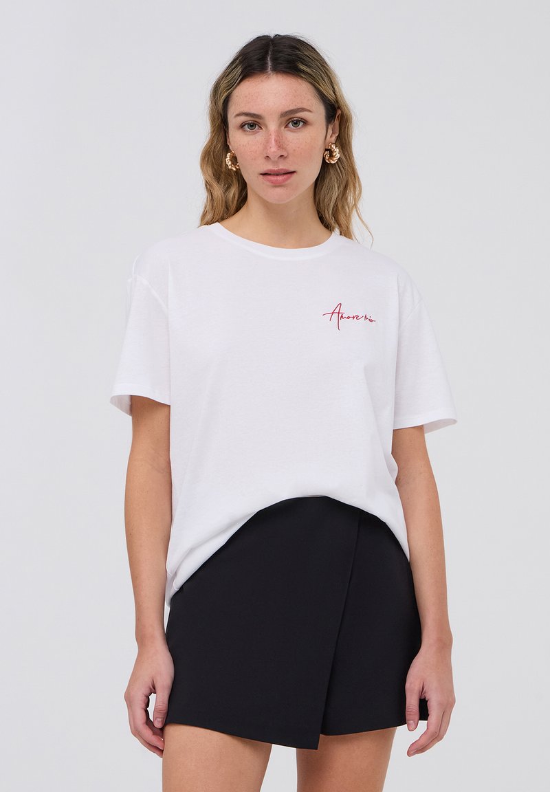 T-shirt bianco in cotone a maniche corte, con un logo rosso in corsivo sul lato sinistro del petto. Abbinato a una gonna nera avvolgente.