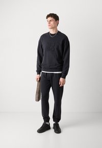 Czarny sweatshirt z wytłoczonym logo "HUGO", w połączeniu z dopasowanymi, zwężającymi się dresami. Model trzyma szaro-skalisty torbę na ramieniu, ma na sobie czarne sneakersy.
