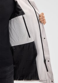 Giacca grigia chiaro con interno nero, dotata di una tasca con cerniera e un'etichetta stampata sulla fodera. Tessuto liscio, design moderno.