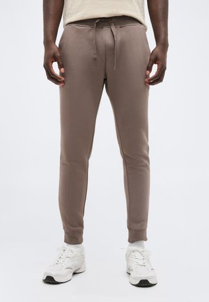 Άνδρας που φοράει ζαχαρί φόρμας με κορδόνι στη μέση και λευκά αθλητικά παπούτσια, στέκεται μπροστά σε απλό ανοιχτό φόντο.