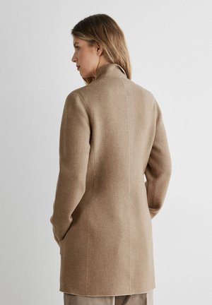 Frau mit langen Haaren, trägt einen mittellangen, hellbraunen Wollmantel mit hohem Kragen, steht mit dem Rücken zur Kamera und den Händen in den Taschen vor einem schlichten Hintergrund.