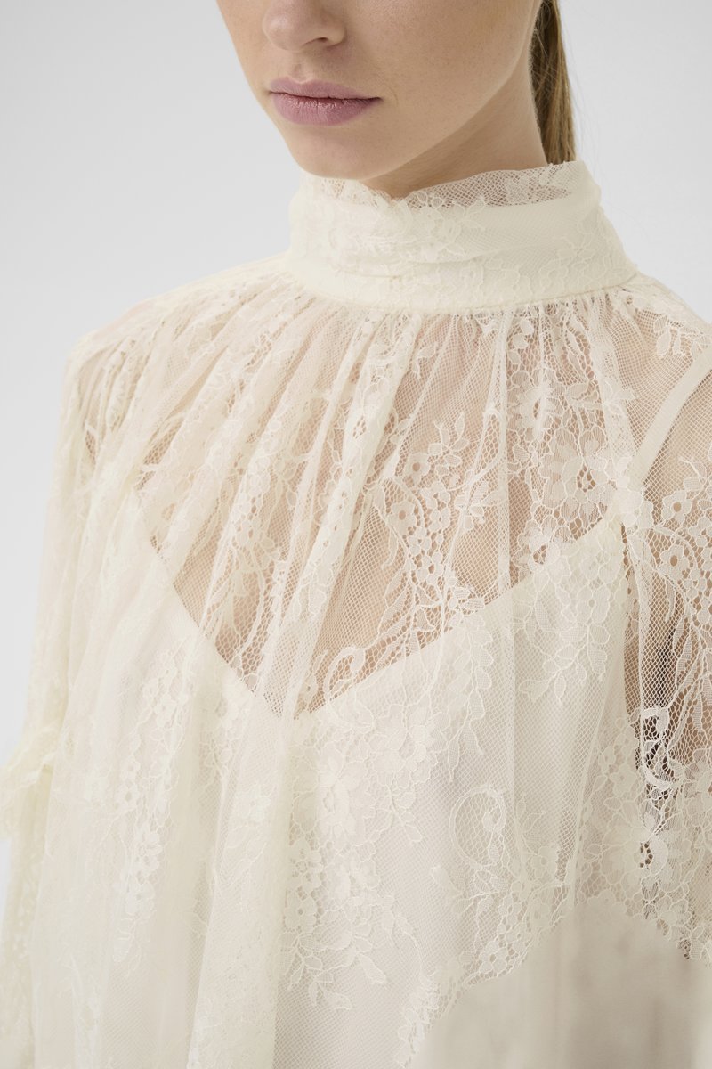 Eine Frau trägt eine hohe, durchsichtige Spitzenbluse mit Blumenmustern über einem weißen Unterhemd, gezeigt von Kinn bis Brust.