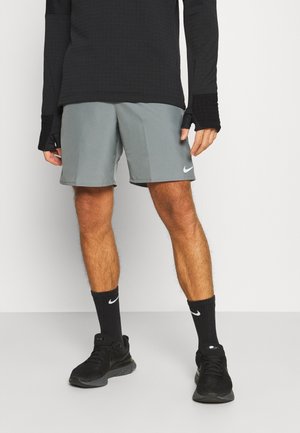 Graue Sportshorts mit glatter Textur, Nike-Logo am linken Saum, kombiniert mit schwarzen Socken und Turnschuhen sowie einem schwarzen Langarmtop.