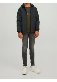 Dziecko w czarnej kurtce z kapturem, oliwkowej koszulce z grafiką, wyblakłych czarnych dżinsach i białych trampkach stoi na jednolitym tle.