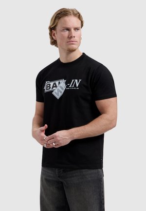 Jonge man met lichtbruin haar, gekleed in een zwart T-shirt met witte "B-ALL-IN" tekst en donkere jeans, kijkt lichtjes opzij.