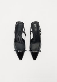 Črne lakirane slingback pete s zašiljenim nosom, DKNY logotipom in elegantno, gladko teksturo. Nastavljiv trak za udobno prileganje.