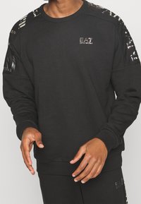Felpa nera a maniche lunghe, con dettagli lucidi neri sulle spalle e un piccolo logo EA7 sul petto. Texture morbida.
