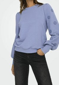 Kvinna iförd en ljuslila sweatshirt med puffärmar och blommig utskärning, i kombination med svarta jeans, med handen i fickan.