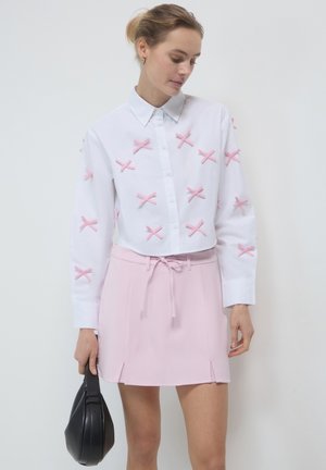 Weißes Hemd mit knöpfbarem Kragen und rosa Stoffschleifen, kombiniert mit einem hellrosa Minirock mit Kordelzugbund und seitlichen Schlitzen. Schwarze Handtasche.