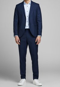 Man som bär en marinblå kostym med ljusblå skjorta och vita sneakers, står mot en enfärgad grå bakgrund.