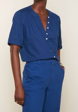 Personne portant une chemise boutonnée à manches courtes bleu cobalt rentrée dans un pantalon taille haute assorti, avec la main dans la poche.