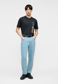 Νεαρός άνδρας που φοράει μαύρο γραφικό μπλουζάκι, ανοιχτόχρωμο μπλε τζιν με μαύρη ζώνη και μαύρα παπούτσια, στέκεται με τα χέρια στις τσέπες.