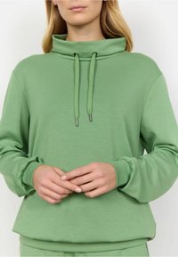 Sudadera verde con cuello alto, cierre con cordones y mangas largas. Textura suave y puños con acentos elásticos.