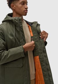Chaqueta verde oliva para hombre con capucha, que presenta un forro aislante naranja y un cierre frontal con cremallera, junto con bolsillos exteriores y cordones ajustables.