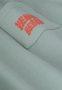 Tissu bleu clair avec une texture côtelée, portant une étiquette affichant « VENICE BEACH » en lettres orange en gras. Le matériau semble doux.