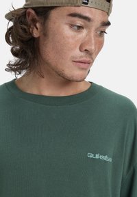 Jonge man met krullend haar, die een beige pet achterstevoren draagt, een diamanten oorbel heeft en een donkergroene Quiksilver-shirt draagt, kijkt naar beneden naar links.
