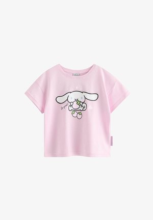 Ung pige står med hånden i lommen, iført en lyserød T-shirt med hundemotiv, blå vidbenede jeans og hvide sneakers.