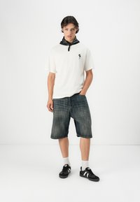 Bela kratka majica s kratkimi rokavi z majhnim črnim dizajnom, v kombinaciji s temnimi jeans kratkimi hlačami do kolen in črnimi supergami z belimi poudarki.
