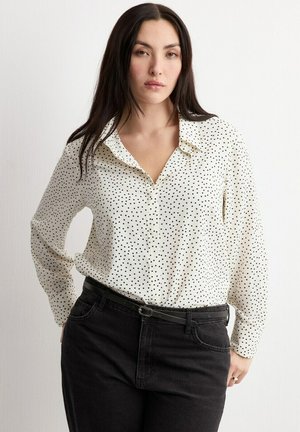 Femme aux longs cheveux noirs portant un chemisier blanc à manches longues à pois noirs, rentré dans un jean noir avec une ceinture noire.