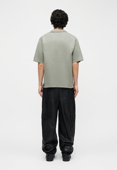 Άνδρας με σγουρά σκούρα μαλλιά, φοράει γκρι κοντομάνικο πουκάμισο και χαλαρό μαύρο παντελόνι cargo, στέκεται με την πλάτη προς το φακό σε απλό φόντο.