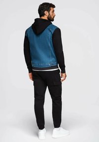 Giacca di jeans con cappuccio, blu davanti, maniche lunghe nere, abbinata a pantaloni cargo neri e sneakers bianche. Caratteristiche includono tasche e polsini elasticizzati.