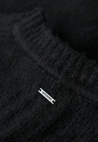 Čierny pletený sveter s rebrovaným výstrihom a malou striebornou nálepkou s nápisom "SUPERDRY" pripevnenou na látke.