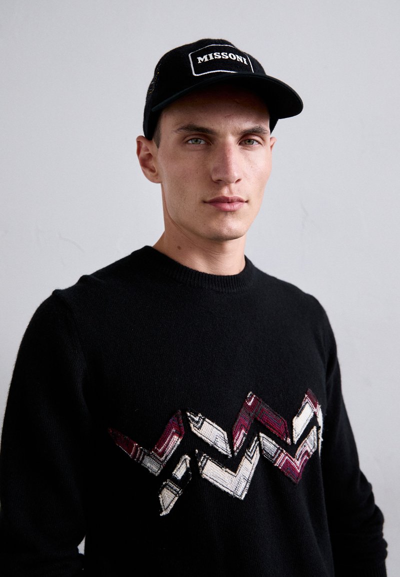 Schwarzer Strickpullover mit einem mehrfarbigen Zickzackmuster auf der Brust, kombiniert mit einer schwarzen Kappe mit dem "MISSONI"-Logo.