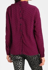 Blouse violette boutonnée avec un motif à pois texturé, dotée d'un col montant et d'une fermeture à boutons dans le dos, associée à un pantalon à imprimé animalier.