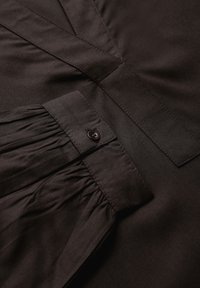 Tissu marron foncé avec une texture lisse. Présente un poignet plissé avec un détail de bouton et un design propre et structuré.