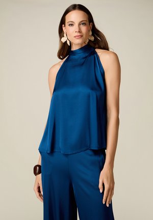 Donna con lunghi capelli castani che indossa un top in raso blu senza maniche con collo halter e pantaloni larghi abbinati, orecchini beige floreali e un braccialetto scuro.