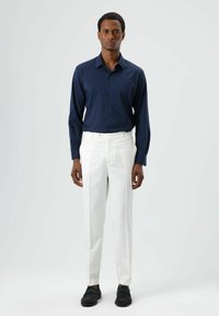 Chemise bleu marine à boutons, pantalon blanc sur mesure et chaussures noires. La chemise a des manches longues ; le pantalon est doté d'une fermeture éclair et de plis.