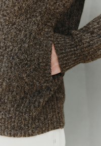 Bruine gebreide trui met textuur en een grote zijzak, voorzien van geribbelde manchetten en zoom. De stof oogt warm en knus.