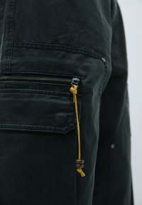 Crne cargo hlače s patentnim zatvaračem, žutom vezicom s dekorativnom kuglicom i strukturiranom tkaninom. Šivani detalji duž šavova.