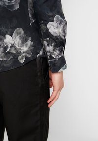 Camicia nera con bottoni, caratterizzata da un motivo a rose in tonalità di grigio e nero, con una texture liscia e maniche lunghe. Orlo frontale visibile.