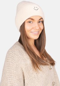 Bonnet en maille côtelée beige orné d'une broderie de visage souriant noir. Porté avec un pull épais de couleur claire.
