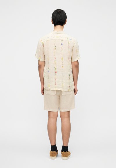 Πουκάμισο από λινό σε ανοιχτό μπεζ χρώμα με κοντά μανίκια και κεντήματα λουλουδιών, συνδυασμένο με μπεζ σορτς και καφέ παπούτσια, από την πίσω όψη.