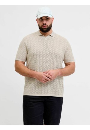 Uomo che indossa una polo beige a maniche corte con trama, pantaloncini neri e berretto azzurro, in piedi con le mani giunte davanti a uno sfondo uniforme.